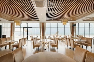Un comedor amplio con mesas y sillas redondas, ventanas grandes y vista al mar.