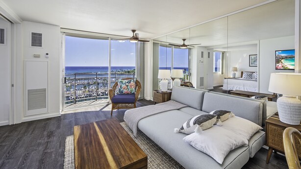 Amplia habitación con una cama grande, un sofá, una mesa de centro de madera y un balcón con vista al océano y al paisaje urbano.