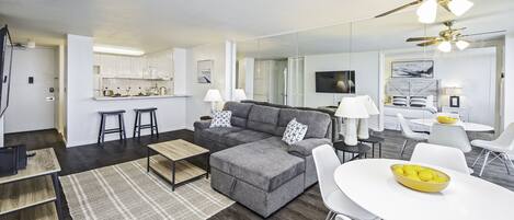 Ein modernes Wohnzimmer mit einem grauen Ecksofa, einem weißen Esstisch und einem Deckenventilator.