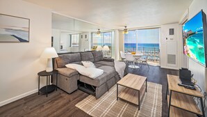 A modern living room with a gray sectional sofa, a small coffee table, a TV, and a view of the ocean through large windows. - Ocean-Marina Yacht Views Condo Steps to the Beach w/Full Kitchen & Free WiFi (Honolulu)