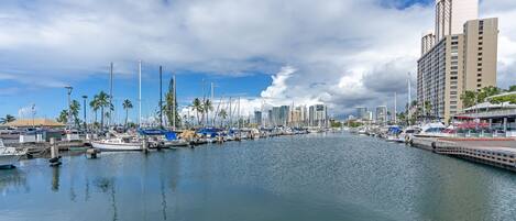 A marina with boats docked, a waterfront promenade, and modern buildings in the background.