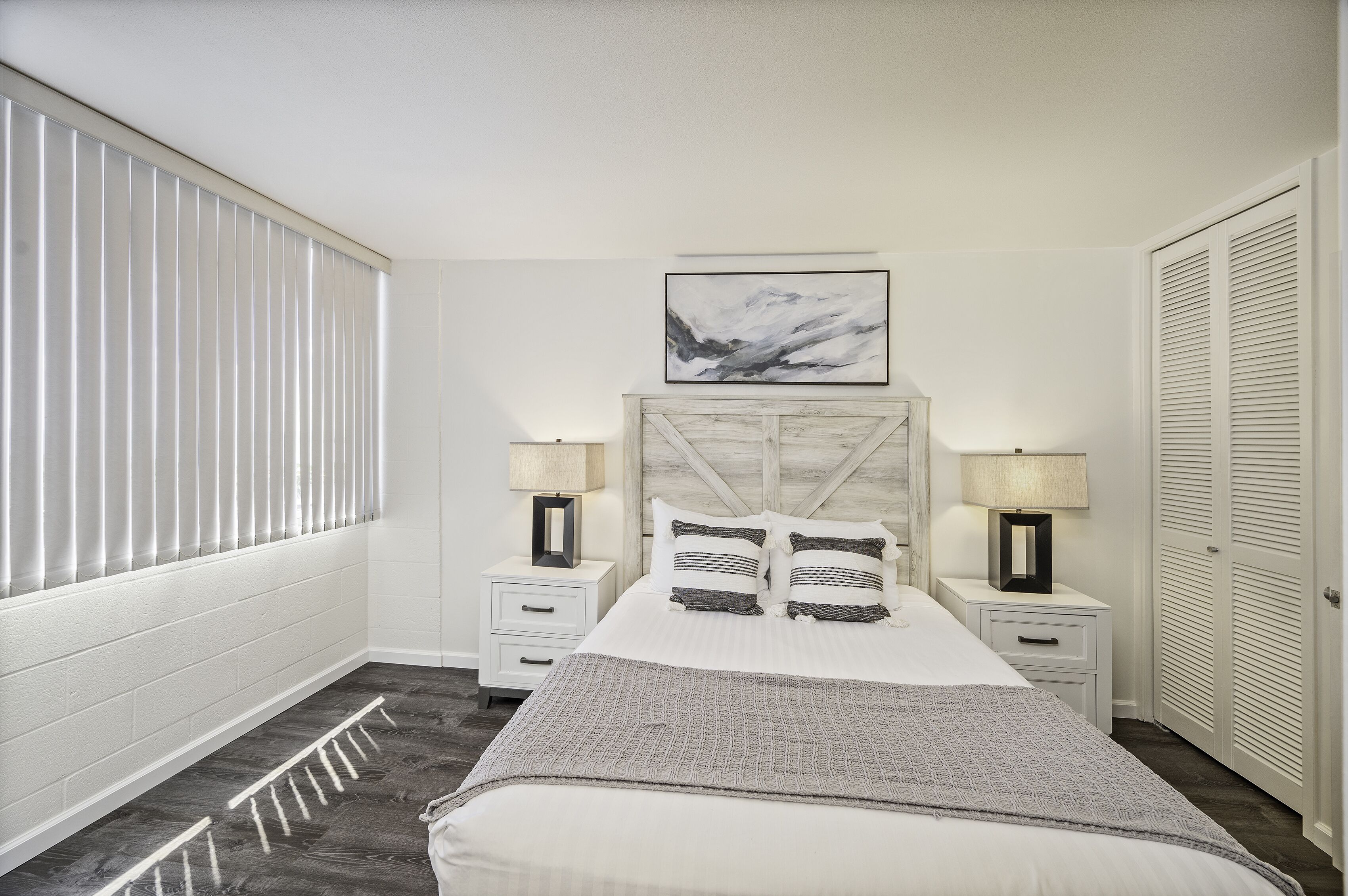 A bedroom with a bed, two bedside tables with lamps, a headboard, a window with blinds, and a door with louvered shutters.