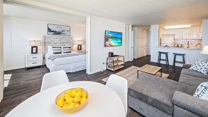 A modern, well-lit living room with a gray sofa, a white dining table with a bowl of fruit, a bed with white bedding, and a TV mounted on the wall. - Ocean-Marina Yacht Views Condo Steps to the Beach w/Full Kitchen & Free WiFi (Honolulu)