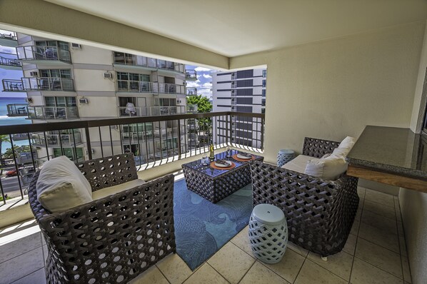 A balcony with wicker furniture, a small table, and a view of the ocean and buildings.