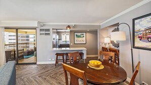 Un área de comedor con una mesa redonda, sillas y un cuenco de fruta. Se observa una cocina al fondo, con un refrigerador y un lavaplatos.