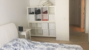 Ett sovrum med en säng, en vit garderob med flera fack, ett sängbord med en lampa och en dörr som leder till ett annat rum.