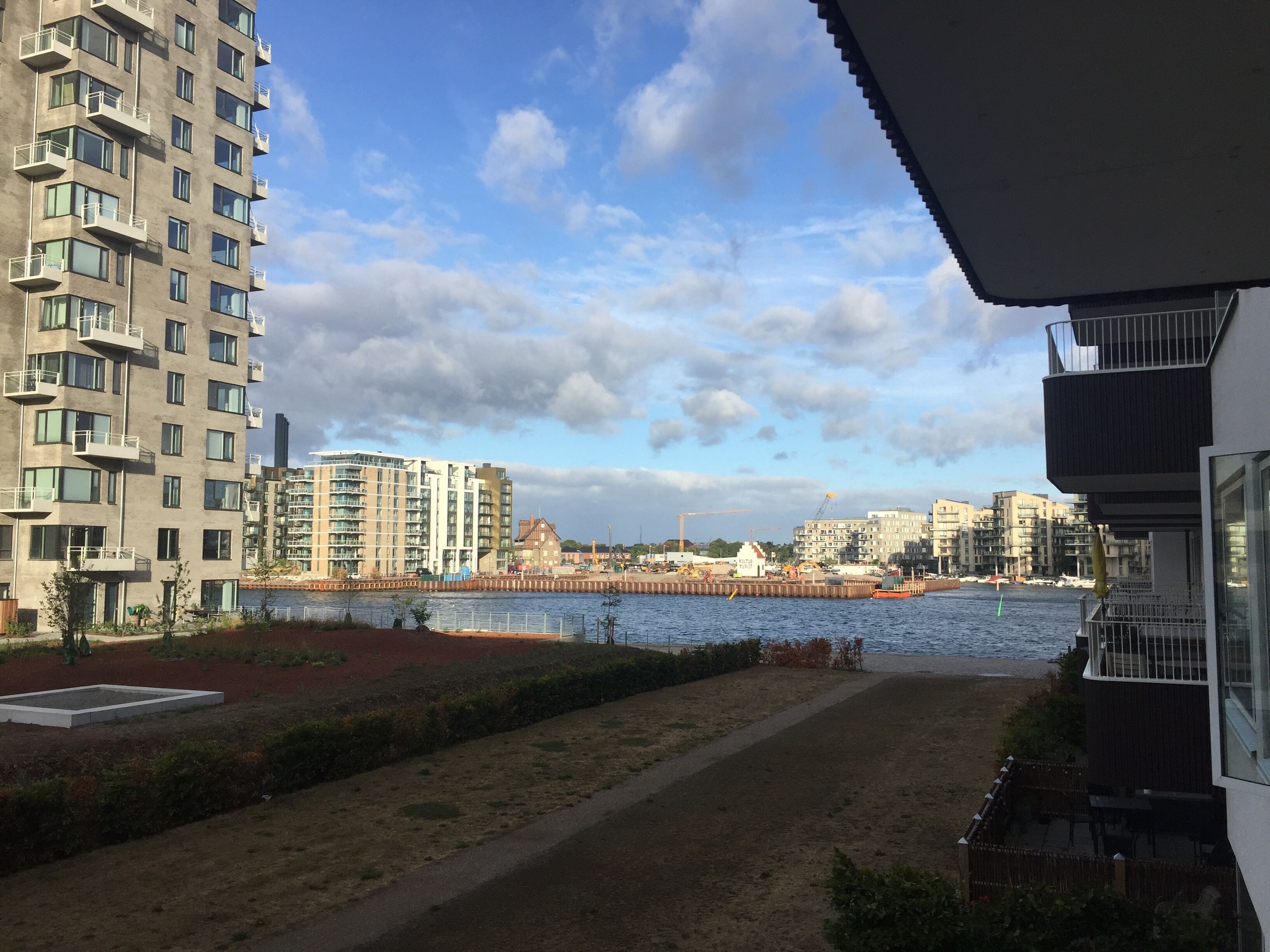 A view of a riverside promenade with modern buildings, a clear sky, and a calm water body.