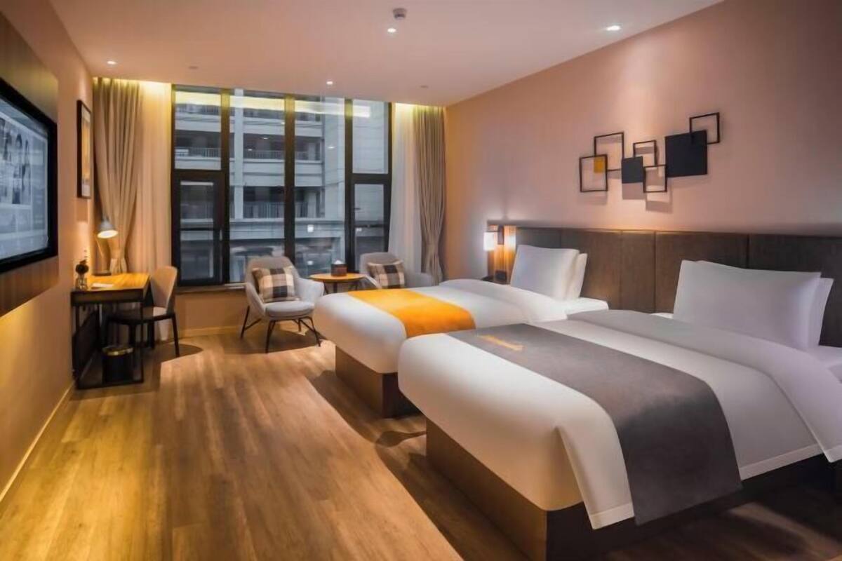 Une chambre d’hôtel avec deux lits, un bureau, une chaise, une petite table et une grande fenêtre avec des rideaux.