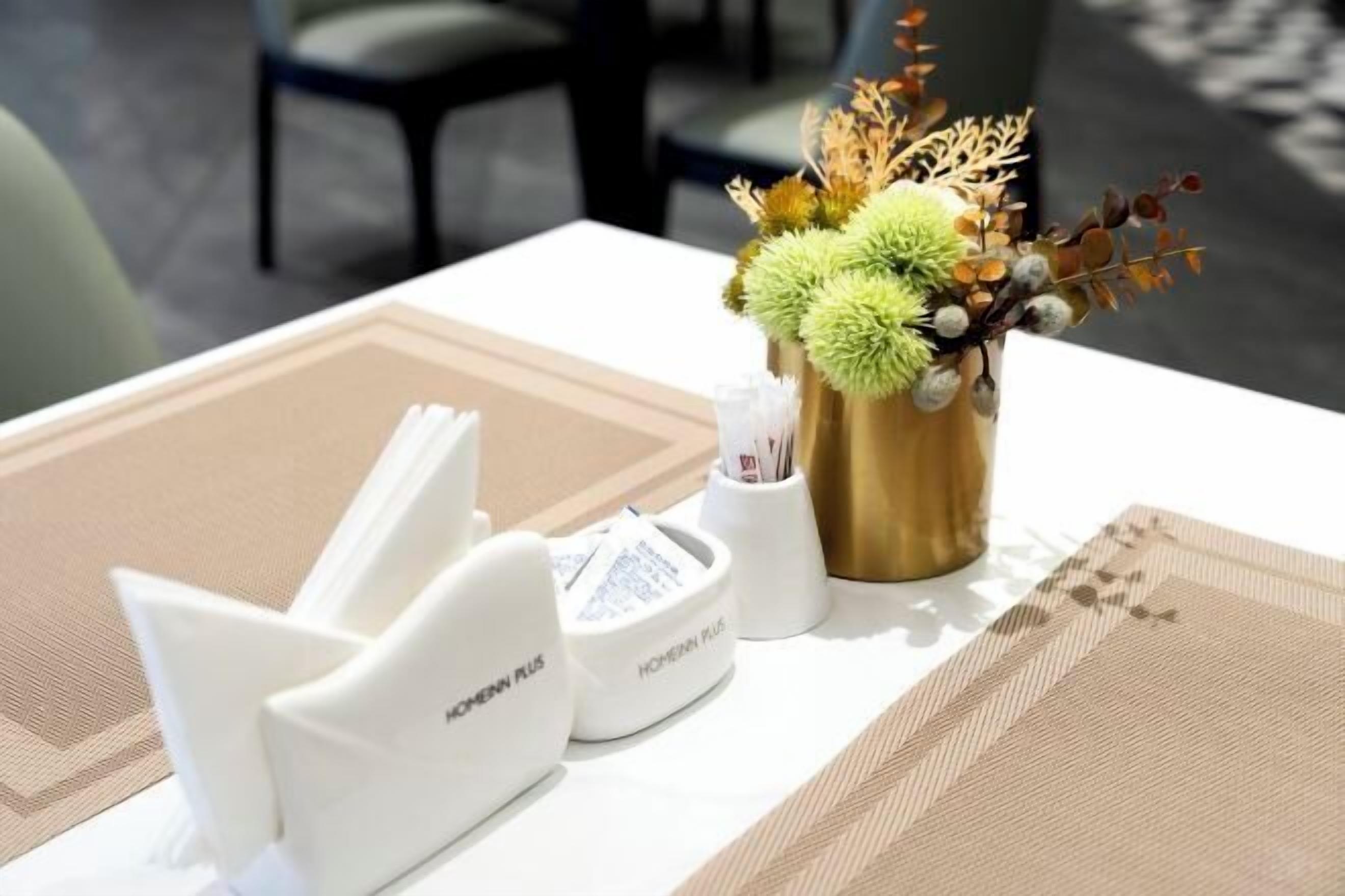 Une table avec un set de table blanc, un vase doré contenant des fleurs séchées et un objet blanc portant l’inscription « HOMANN PLUS ».