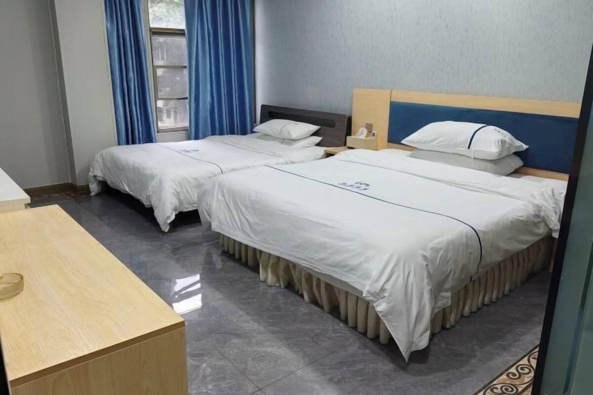 Een hotelkamer met twee enkele bedden, een houten bureau, een raam met blauwe gordijnen en een betegelde vloer.
