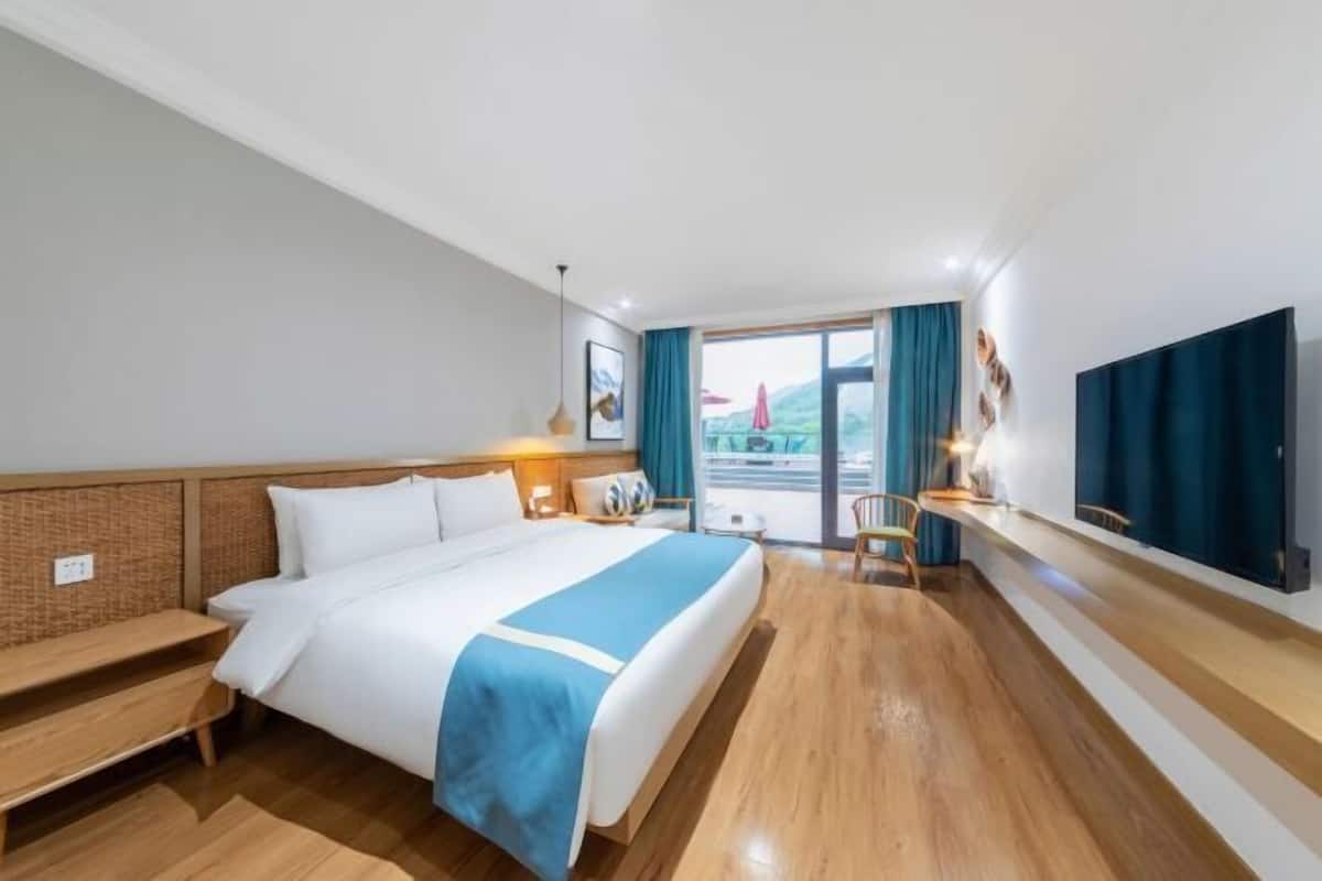 A hotel room with a bed, bedside table, lamp, and a view of the outside.