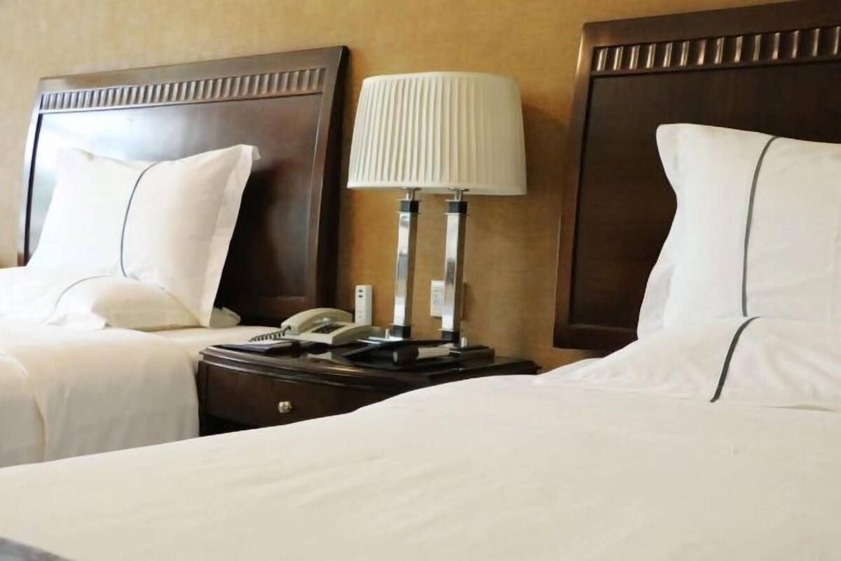 A hotel room with two beds, a nightstand with a lamp, a phone, and a wall-mounted device.