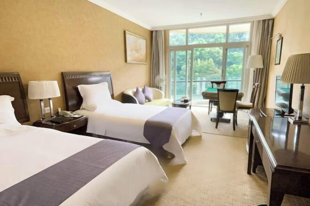 A hotel room with two beds, a desk, a chair, and a large window offering a view of greenery.