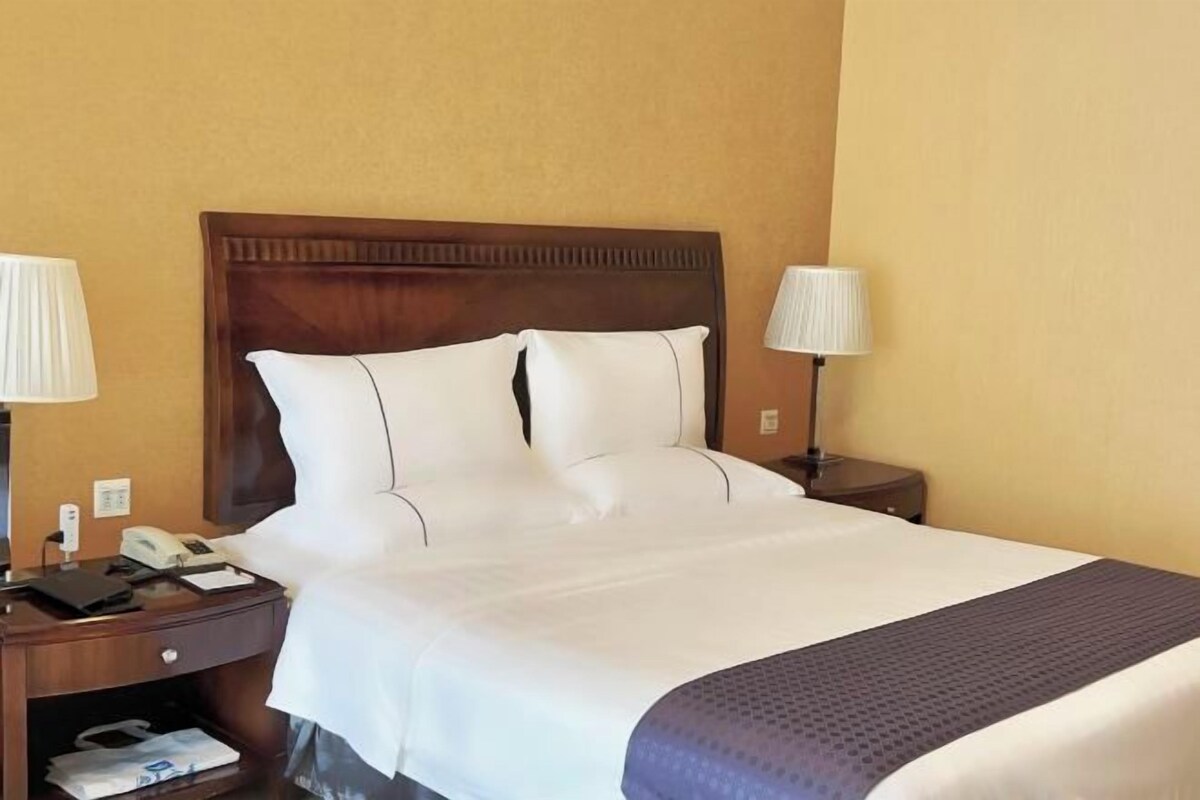 A hotel room with a wooden headboard, white bedding, a telephone, a lamp, and a nightstand with a bag.