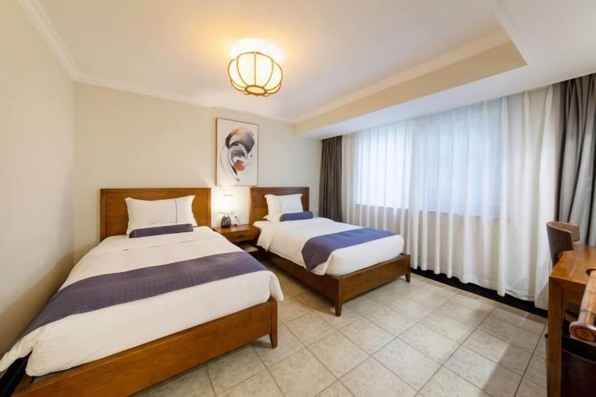 A hotel room with two beds, a nightstand, a lamp, a painting on the wall, and a window with curtains.