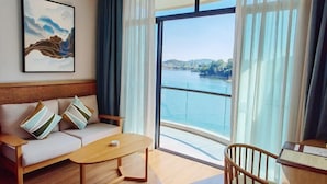 A room with a sofa, a wooden coffee table, a chair, a large window with a view of a body of water, and a framed artwork on the wall. - Sky & Sea Resort (Chongqing)