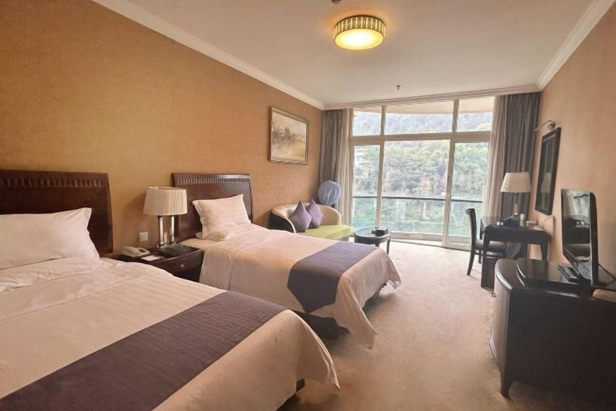 A hotel room with two beds, a desk, a chair, a TV, and a large window with a view of trees.