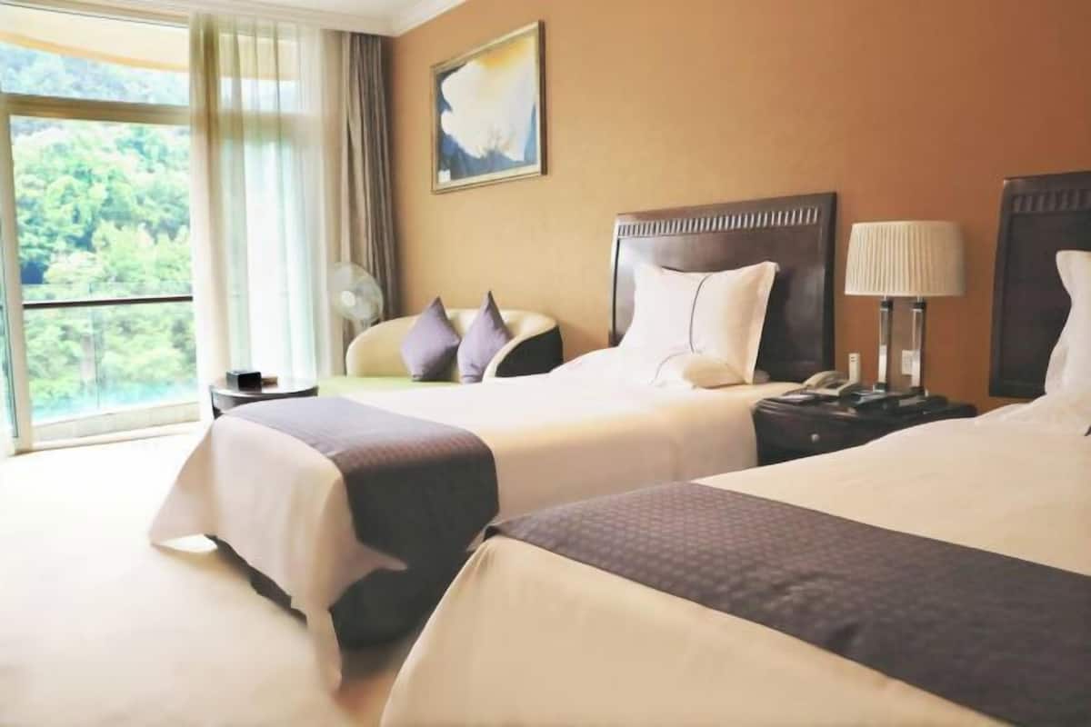 A hotel room with two beds, a large window, a painting on the wall, and a lamp on the nightstand.
