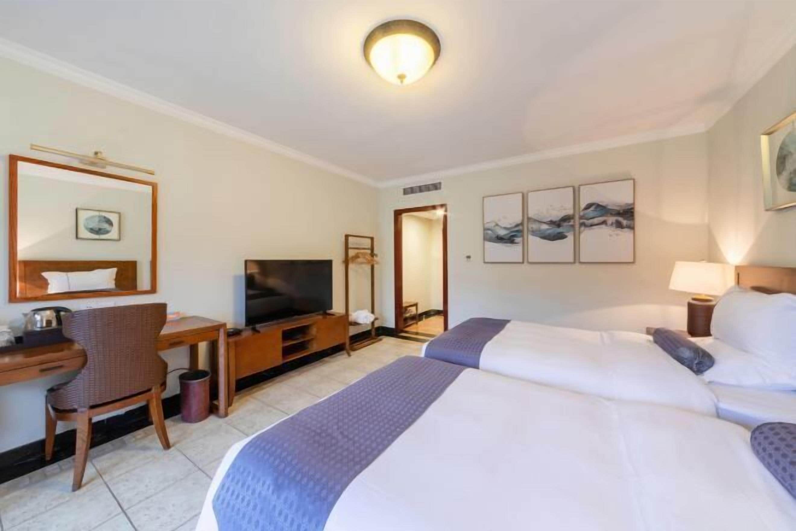 Una habitación de hotel con dos camas, un escritorio con televisor, una silla y obras de arte en las paredes.