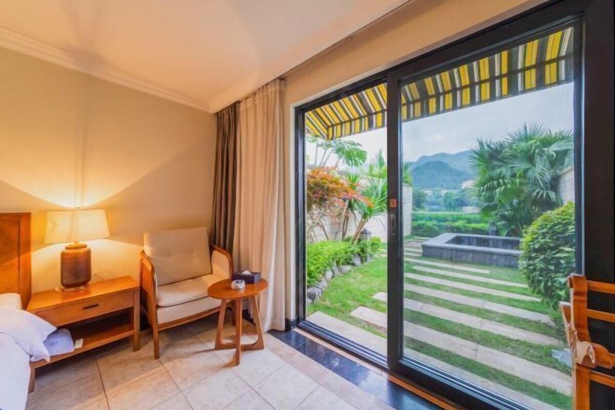 A hotel room with a bed, a bedside table with a lamp, a sofa, a small table, and a large sliding glass door leading to a garden.