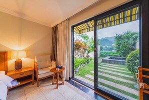 A hotel room with a bed, a bedside table with a lamp, a sofa, a small table, and a large sliding glass door leading to a garden. - Sky Sea Resort (Chongqing)