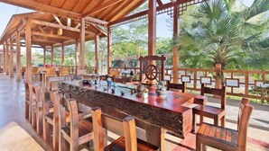 An outdoor dining area with wooden furniture and a large table, surrounded by a wooden pergola with a view of palm trees. - Lingshui Sancai Business Hotel (Lingshui)