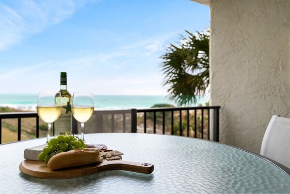 A table with a bottle of wine, two glasses, grapes, a baguette, and crackers on a glass table with a view of the ocean.