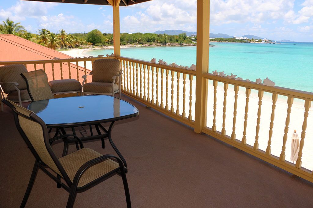 A covered outdoor seating area with a table and chairs, overlooking a beach and turquoise waters.