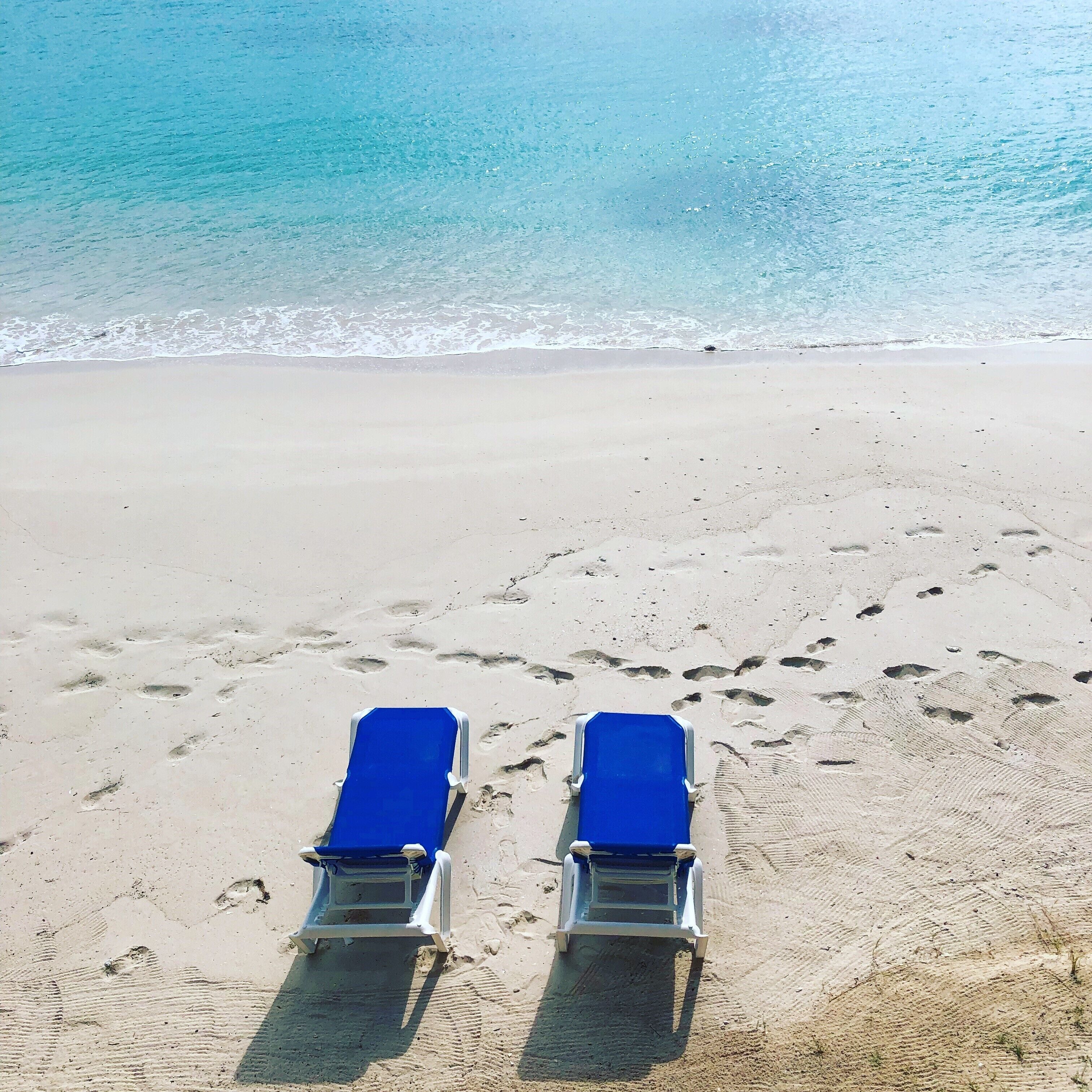 Duas cadeiras azuis na areia de uma praia, com pegadas que vão até a água.