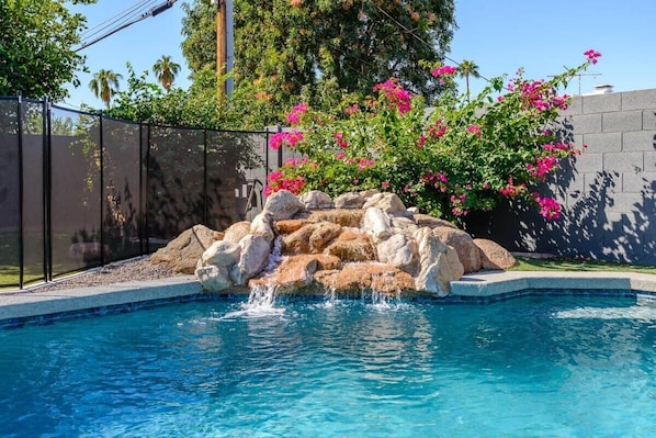 A swimming pool with a rock feature and pink flowers.