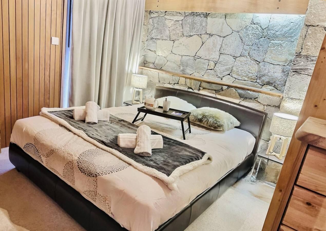 Un dormitorio con pared de piedra, cama con cabecera, mesita de noche y velador.
