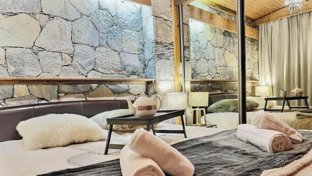 Quarto com parede de pedra, cama com toalhas dobradas, mesa pequena com bule e janela com cortinas.