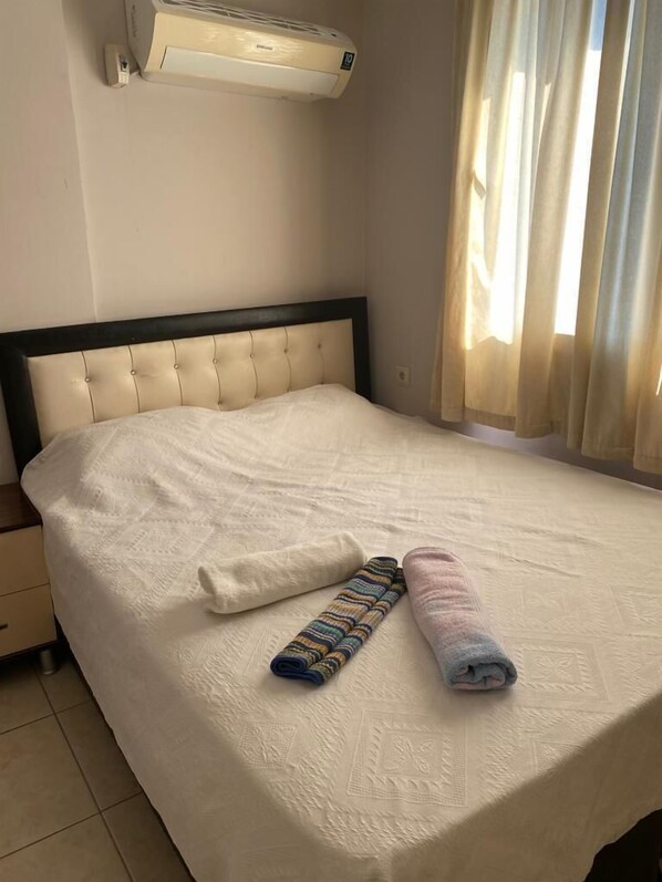 A neatly made bed with a white duvet, two rolled towels, and a Samsung air conditioner above it.
