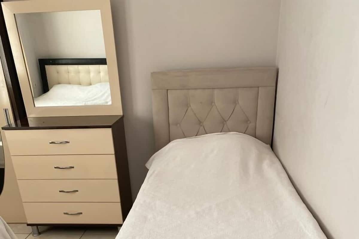 Un petit lit bien fait, recouvert d’une courtepointe blanche, dans une pièce où se trouvent une commode beige et un miroir.
