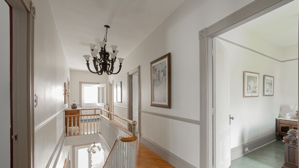 Ein geräumiger Flur mit Holzboden, weißen Wänden und einem Kronleuchter. Eine Treppe mit weißen Geländern führt auf eine obere Ebene.