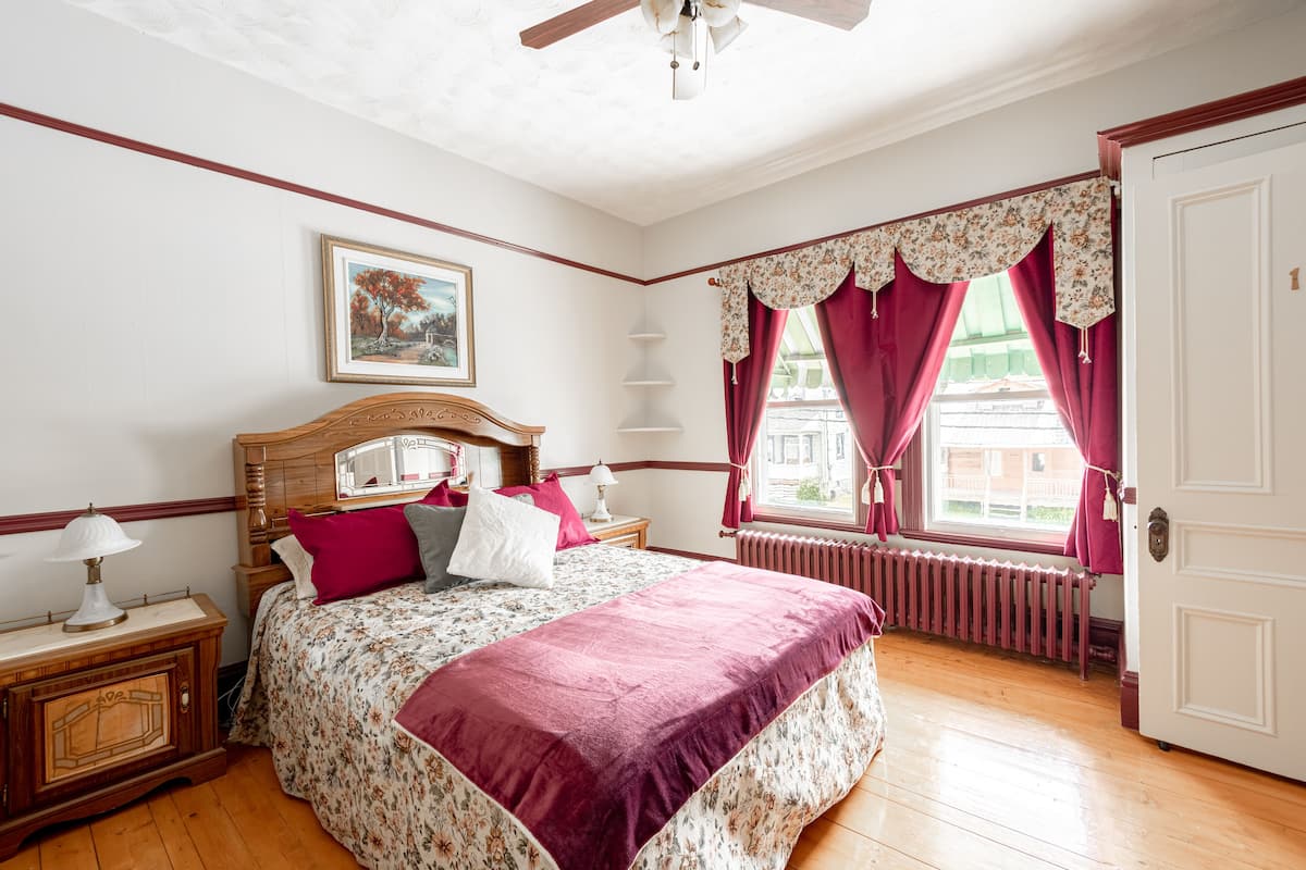 A bedroom with a wooden bed, a nightstand, a lamp, a radiator, and a window with curtains.