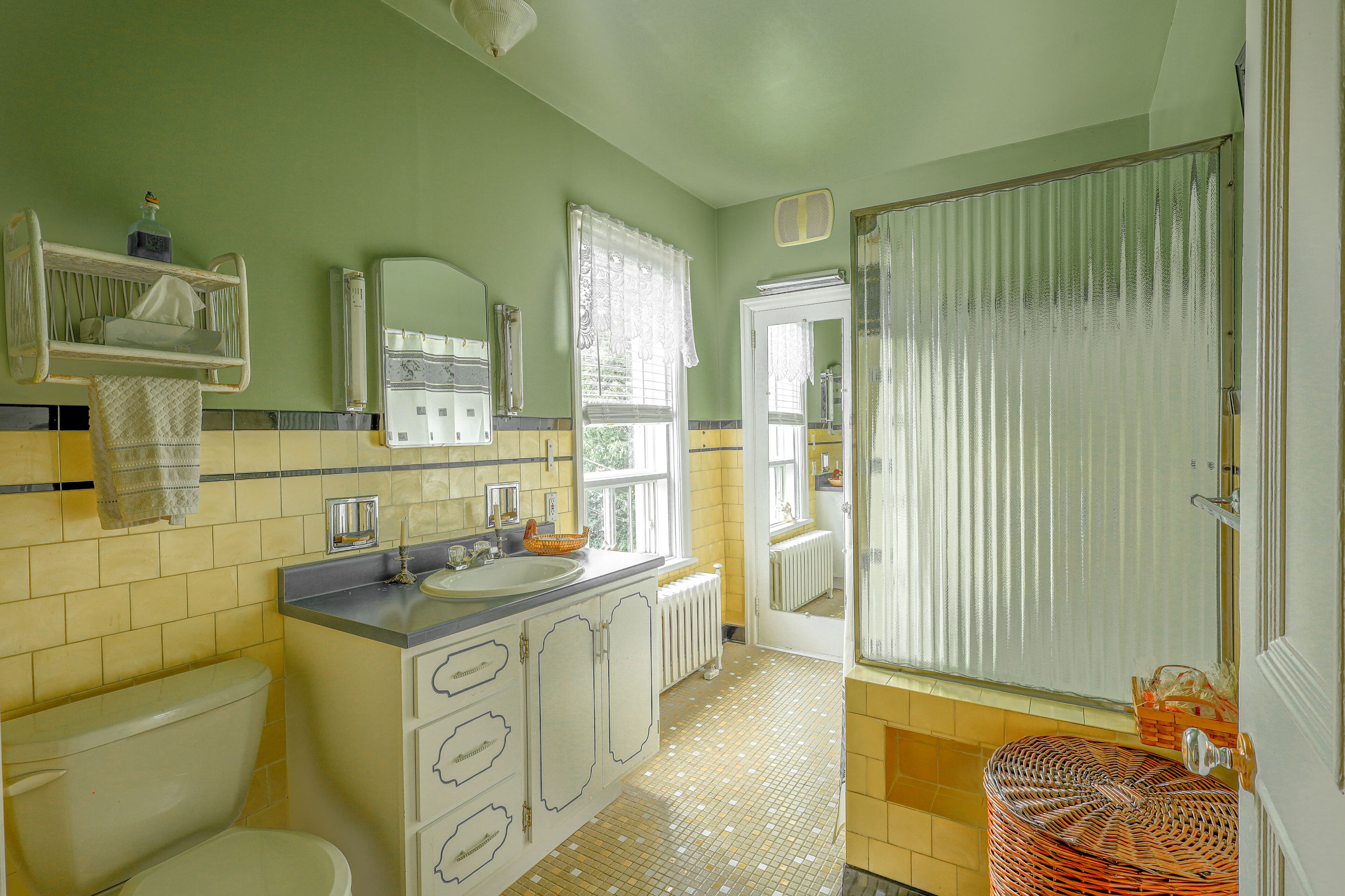 Un baño con inodoro, lavabo y espejo. Hay una cesta en el suelo y una ventana con cortinas.
