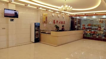 A hotel reception area with a reception desk, a television mounted on the wall, and a chandelier.