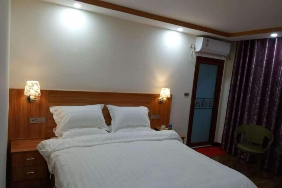 Habitación de hotel con cabecera de madera, dos lámparas de noche, cama con ropa blanca, mesita de noche, unidad de aire acondicionado, puerta con cortina y una silla.