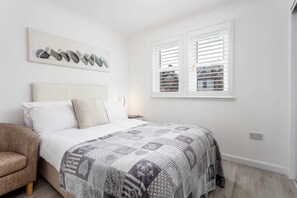 Un dormitorio con cama, una silla, una ventana con persianas y una obra de arte colgada en la pared.