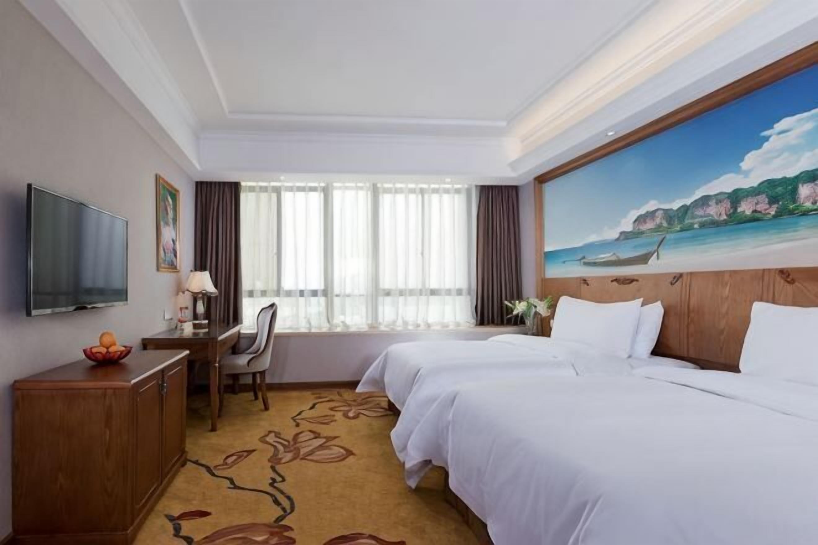 A hotel room with a bed, desk, TV, and a painting of a beach scene.