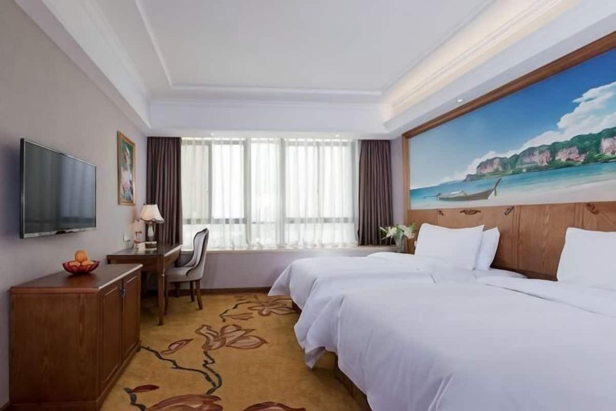 A hotel room with a bed, desk, TV, and a painting of a beach scene.