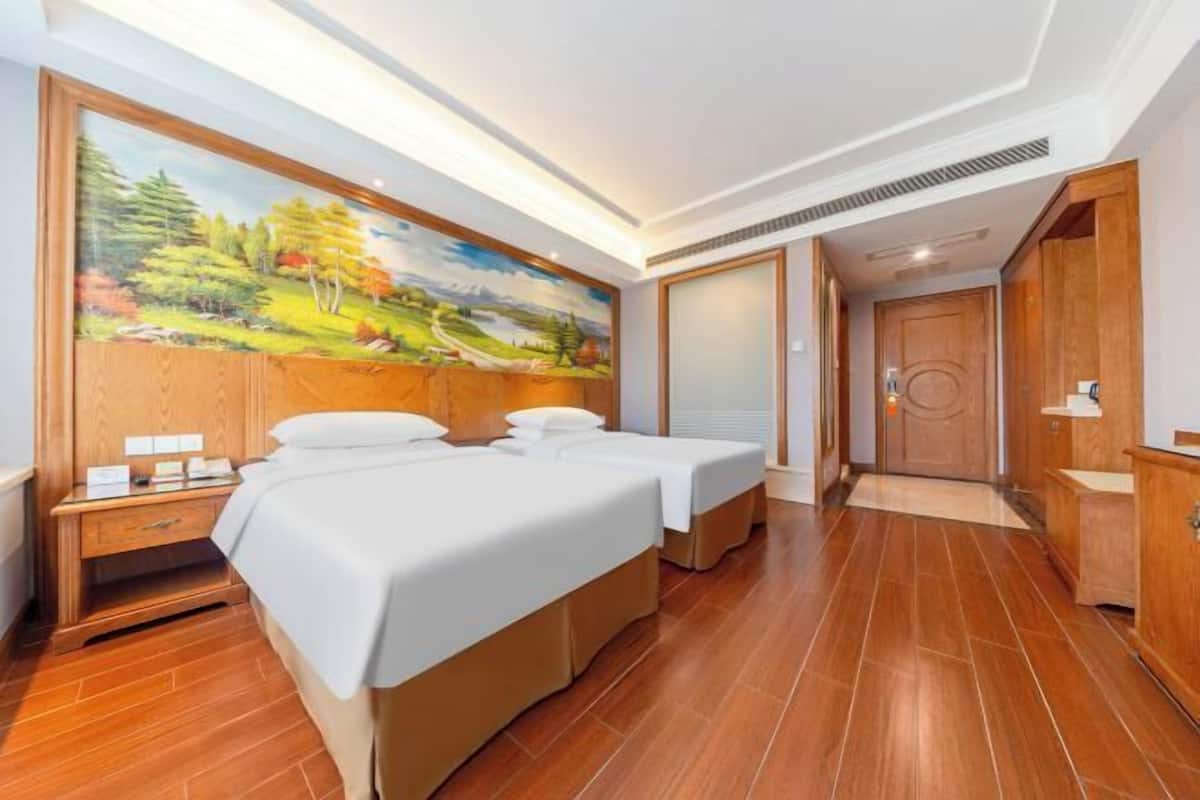 A hotel room with two beds, a wooden headboard, a painting of a landscape, a wooden floor, and a wooden door.