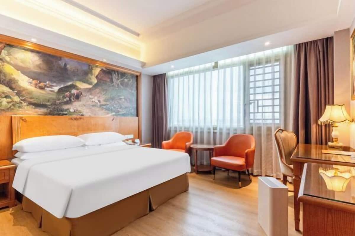 A hotel room with a bed, a desk, two chairs, a painting on the wall, and a window with curtains.