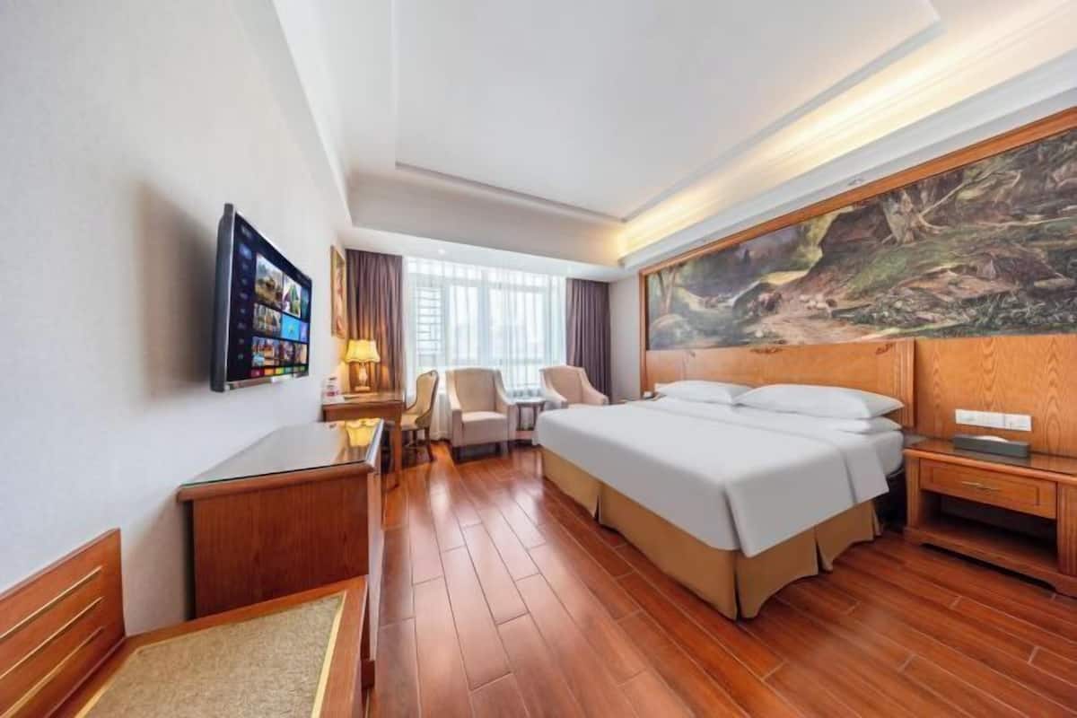 A hotel room with a bed, desk, chair, TV, and artwork on the wall.