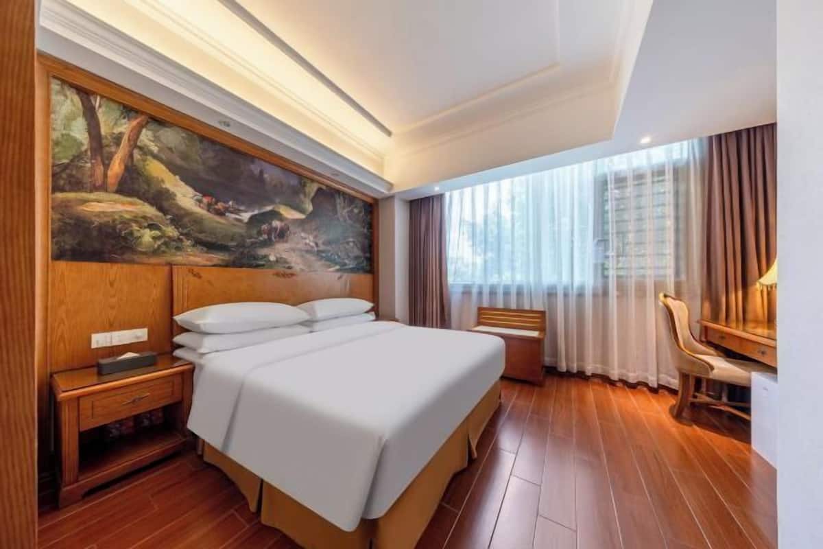 A hotel room with a large bed, a nightstand, a desk with a chair, and a painting on the wall.