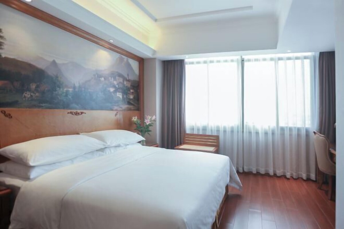 Quarto de hotel com uma cama grande, cabeceira de madeira, pintura de uma paisagem montanhosa, escrivaninha com cadeira e uma janela ampla com cortinas.