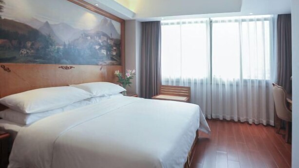 Quarto de hotel com uma cama grande, cabeceira de madeira, pintura de uma paisagem montanhosa, escrivaninha com cadeira e uma janela ampla com cortinas.