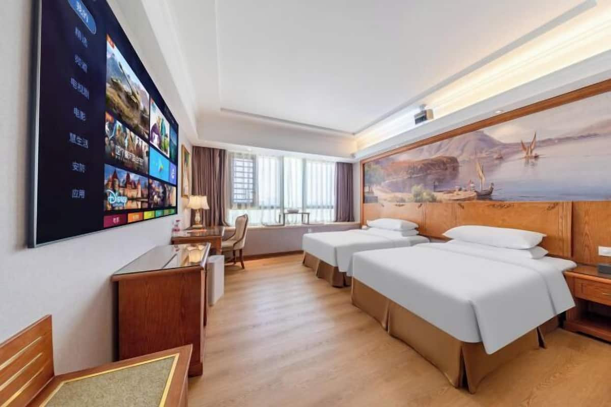 A hotel room with two beds, a desk, a chair, a TV mounted on the wall, and a painting of a seascape.