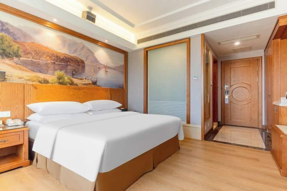A hotel room with a large bed, wooden headboard, a painting of a desert landscape, a wooden wardrobe, and a door leading to another room.